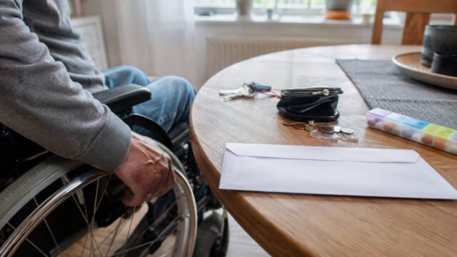Rollstuhlfahrer neben Tisch mit Briefumschlag, Geldbörse, Münzen und Medikamentenbox