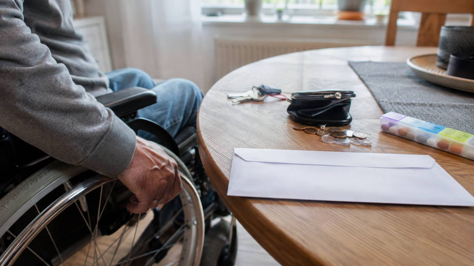 Rollstuhlfahrer neben Tisch mit Briefumschlag, Geldbörse, Münzen und Medikamentenbox