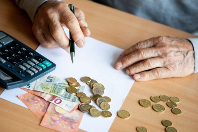 Rentner berechnet mit Taschenrechner und Stift das Haushaltsbudget, auf dem Tisch liegen Euro-Scheine und Münzen. Symbolbild für Wohngeld, finanzielle Unterstützung und Rentnerhaushalt.